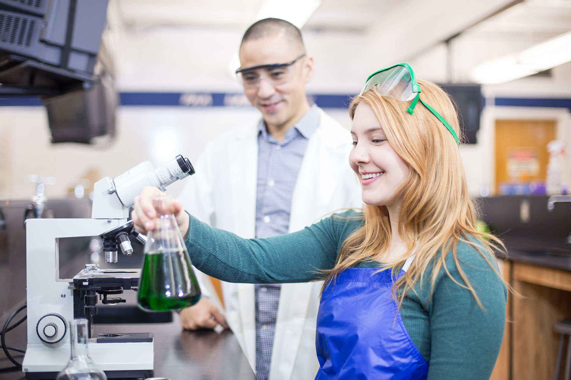Eine schülerin hält einen Glaskolben mit grüner Flüssigkeit hoch, während sie und ein Chemikant lächeln