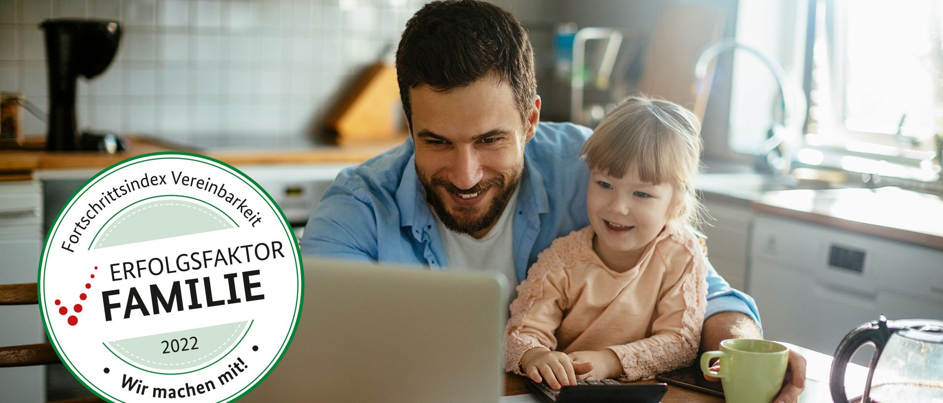 Vater sitzt mit seiner kleinen Tochter am Küchentisch und sie schauen glücklich auf den Laptop vor ihnen.