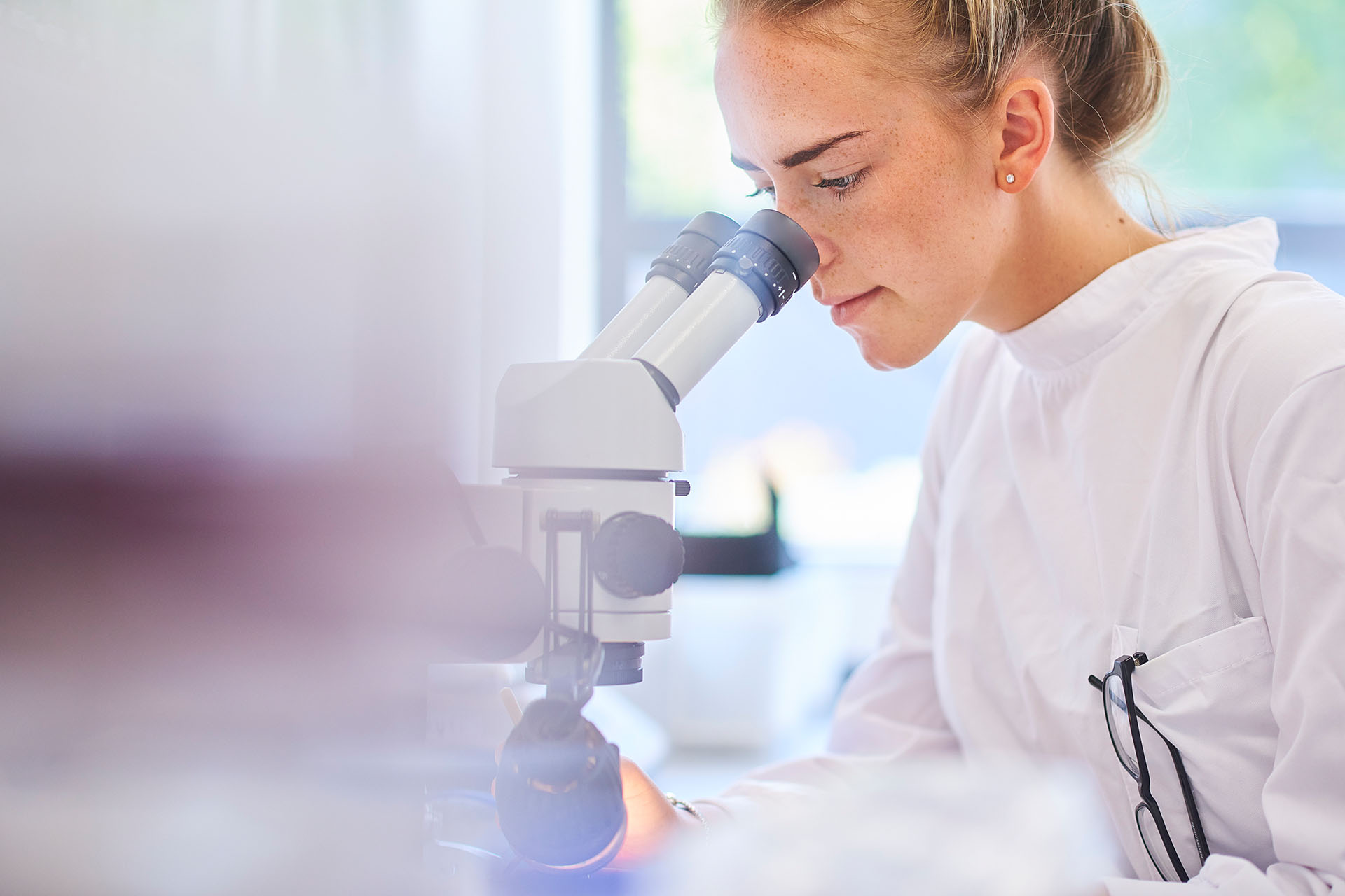 Schülerin in einem weißen Laborkittel schaut durch ein Mikroskop