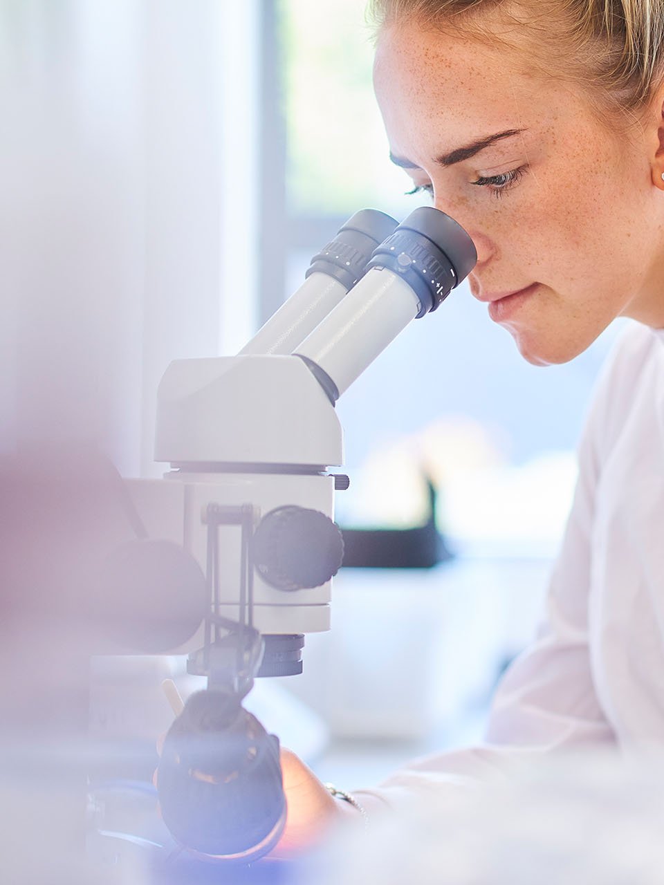 Schülerin in einem weißen Laborkittel schaut durch ein Mikroskop
