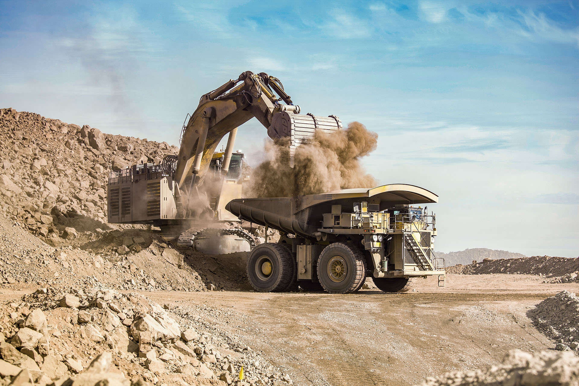 Excavator unloads shovel with dusty soil into large dump truck